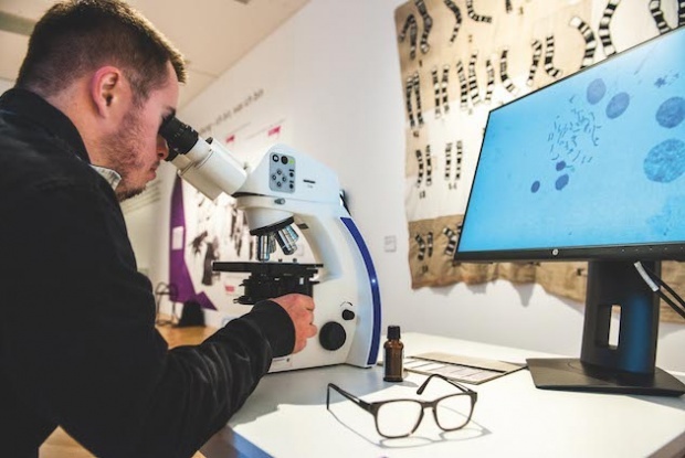 Ein Mann mit Down-Syndrom guckt durch ein Mikroskop. Die Chromosomen kann man vergrößert auf einem Bildschirm sehen.