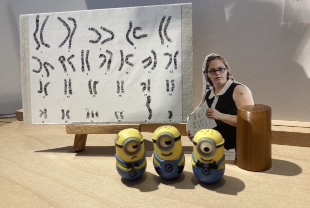 3 Minion-Figuren stehen vor einer Tafel mit Chromosomen, daneben das Foto einer Frau mit Down-Syndrom in schwarz-weißem Kleid.