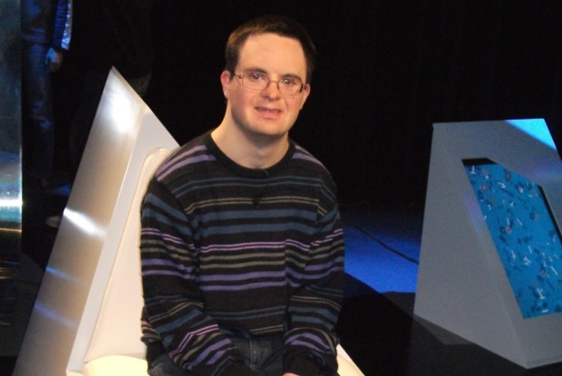 Ein Mann mit Down-Syndrom mit kurzen dunklen Haaren und Brille in einem gemusterten Strickpullover.
