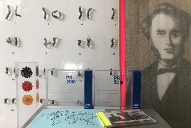 Pokale aus Plexiglas stehen auf einer Metallkiste, im Hintergrund sieht man John Langdon-Down und Chromosomen
