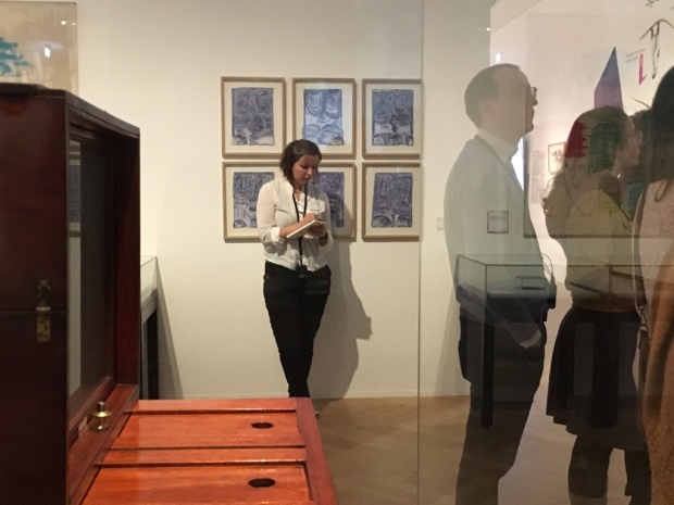 Besucher und Besucherinnen stehen in der Ausstellung, Thea Jacob steht mit etwas Abstand daneben und macht sich Notizen. Hinter ihr sieht man 4 Bilder eines belgischen Künstlers mit Down-Syndrom.