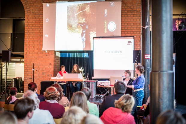 2 Frauen sitzen auf der Bühne, 2 stehen davor. Hinter ihnen ist eine große Projektion zu sehen. Zuschauerinnen und Zuschauer sitzen vor der Bühne.