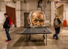 Eine halbe Tischtennisplatte an deren Kopfende ein Portrait des Wissenschaftlers Pawlow montiert ist. 2 Menschen gehen am Kunstwerk vorbei.