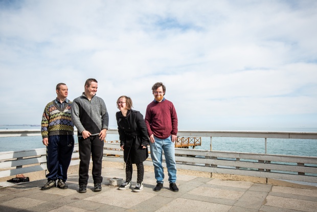 4 Personen mit Down-Syndrom stehen nebeneinander am Meer, davon 3 Männer und eine Frau