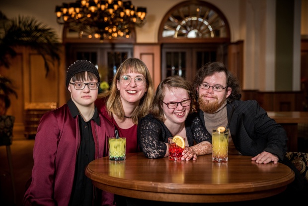 3 Personen und eine Person ohne Down-Syndrom in einer Bar, auf dem Tisch stehen Cocktails. Alle sind elegant gekleidet.
