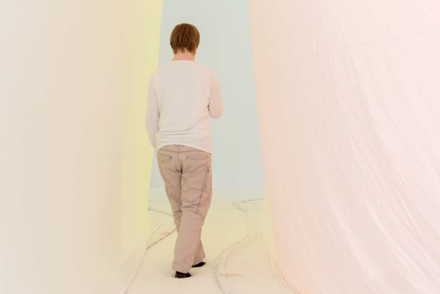 Ein Mann mit Down-Syndrom von hinten fotografiert. Er geht durch einen hellen Tunnel aus Stoff.