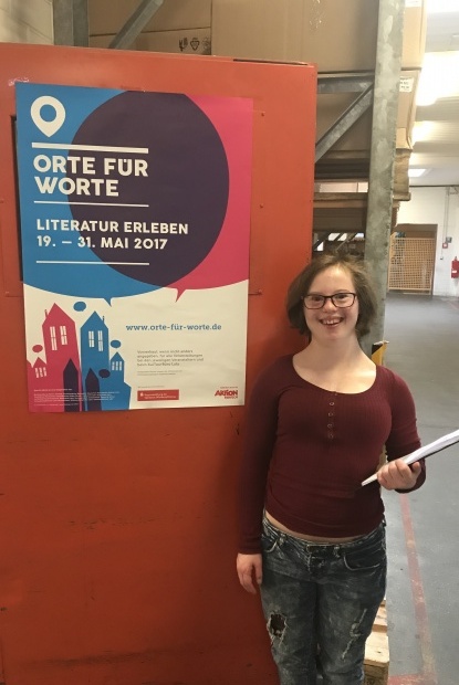 Natalie Dedreux vor einer roten Säule mit  dem Plakat Orte für Worte. Sie hält eine Klemmmappe mit ihren Lesungstexten in der Hand.