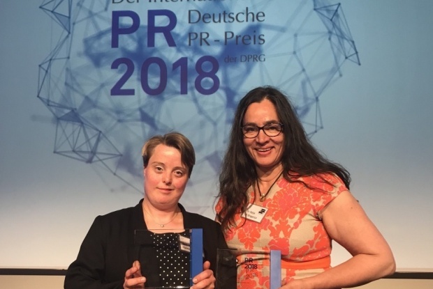 Andrea Halder und Katja de Bragança mit den 2 Trophäen des Deutschen Internationalen PR-Preises: Sie halten 2 Plexiglas-Preise in der Hand, im Hintergrund das Logo der Veranstaltung.