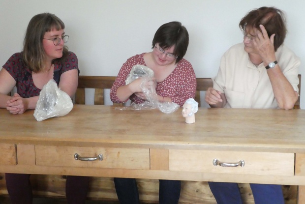 3 Frauen sitzen nebeneinander an einem Holztisch. Eine der Frauen hält eine Keramikfigur in den Händen.