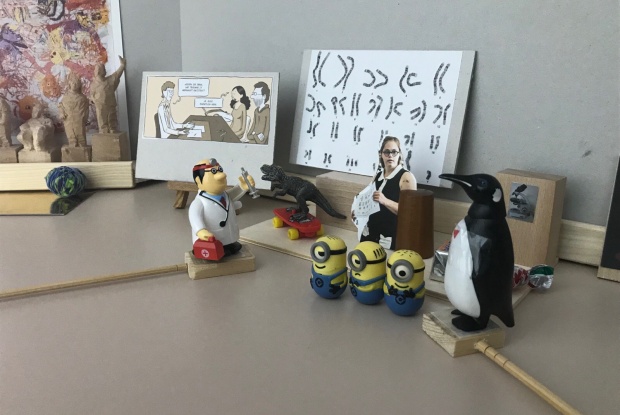 Detailaufnahme: Eine kleine Plastikfigur eines Arztes steht vor einer Abbildung mit Chromosomen. Im Vordergrund 3 Minion-Figuren aus dem Überraschungsei.