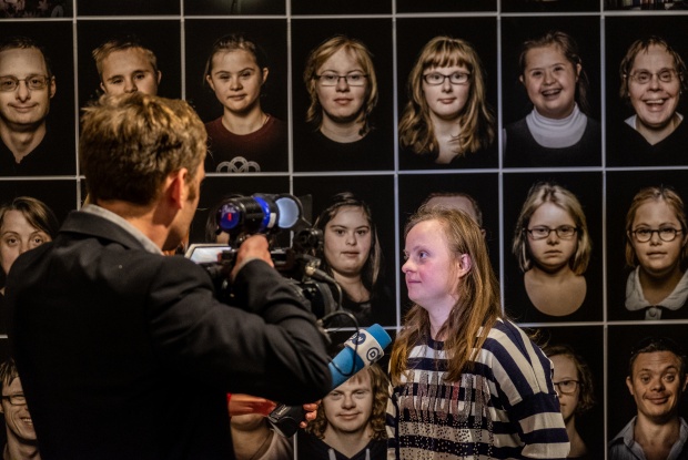 Eine junge Frau mit Down-Syndrom steht vor einer Reihe von Portraitfotos und spricht in das Mikrophon eines Journalisten.