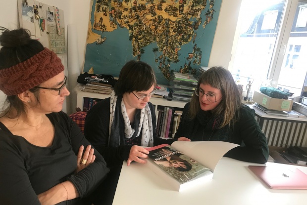 3 Frauen, eine davon mit Down-Syndrom, sitzen an einem Tisch und schauen gemeinsam in ein Buch. Im Hintergrund hängt eine Weltkarte an der Wand.