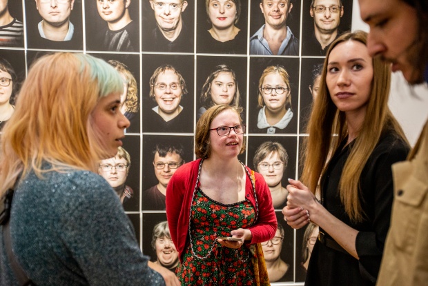 Mehrere Menschen stehen im Gespräch vor einer Wand mit Portraitfotos von Menschen mit Down-Syndrom.