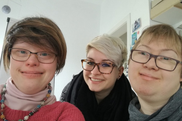 3 Personen machen ein Selfie von sich, zwei Frauen, ein Mann. Zwei der Personen haben das Down-Syndrom. Alle drei tragen eine Brille.
