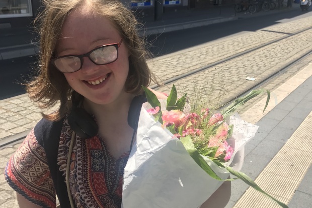 Natalie Dedreux steht in einem gemusterten Oberteil an einer Bahnhaltestelle, lächelt und hält einen Blumenstrauß in der Hand.