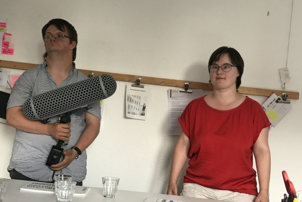 Anna-LisaPlettenberg spricht in die Kamera. Julian Göpel hält ein großes Mikrophon. Hinter beiden sieht man eine Holzleiste, an der Zettel hängen.