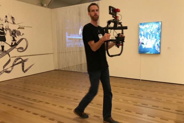Ein sehr großer, dünner Mann geht mit einer Kamera, die in einem tragbaren Stativ hängt, durch die Ausstellungsräume des Zentrum Paul Klee