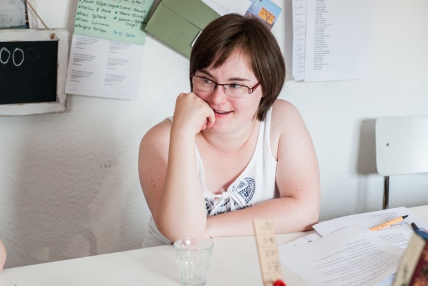 Eine dunkelhaarige Frau mit Down-Syndrom mit Brille und einem hellen Top. Sie sitzt an einem Tisch, auf eine Hand gestützt, und lächelt.
